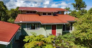 A house with a red metal roof surrounded by trees and greenery under a partly cloudy sky.