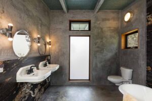 Industrial-style bathroom with concrete walls, two round mirrors, dual sinks, and a toilet. A tall frosted window allows light, and a circular wall light is above one mirror.